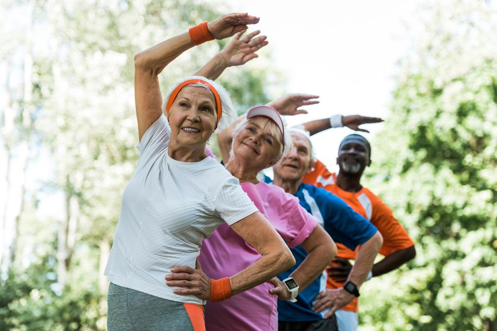 Selektiver Fokus glücklicher multikultureller Senioren, die mit den Händen auf den Hüften stehen, während sie Sport treiben  - Foto, Bild