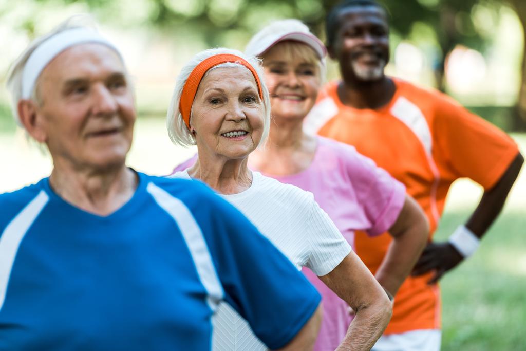 Selektiver Fokus einer fröhlichen Rentnerin, die mit den Händen auf den Hüften steht, mit multikulturellen Rentnern in Sportkleidung  - Foto, Bild