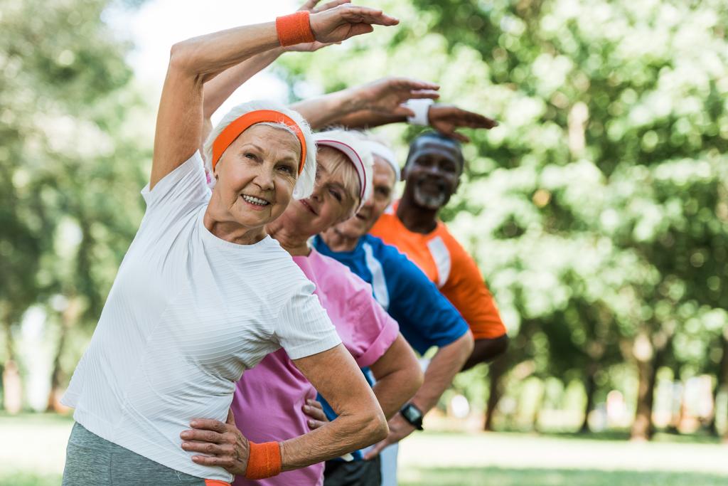 selective focus of cheerful retired woman near multicultural men and woman doing stretching exercise  - Photo, Image