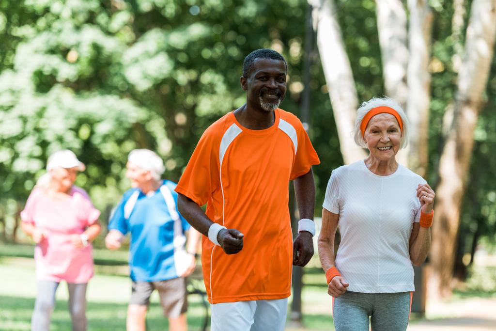 foco seletivo do homem sênior afro-americano e da mulher aposentada feliz exercitando perto de aposentados fora
  - Foto, Imagem