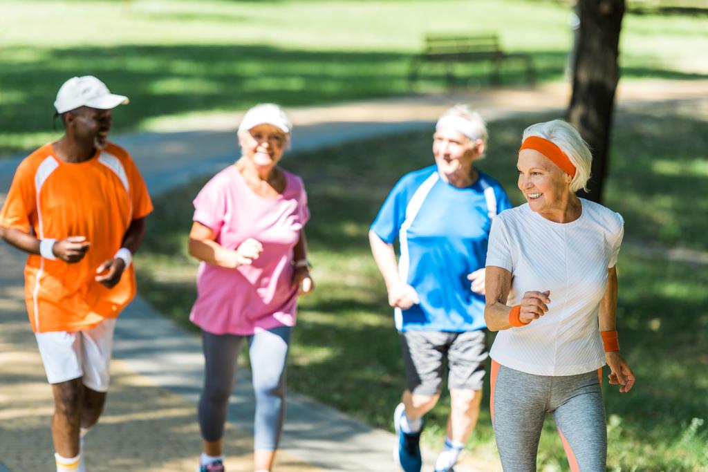 公園で多文化年金受給者と一緒に走っている幸せな退職した女性の選択的な焦点  - 写真・画像