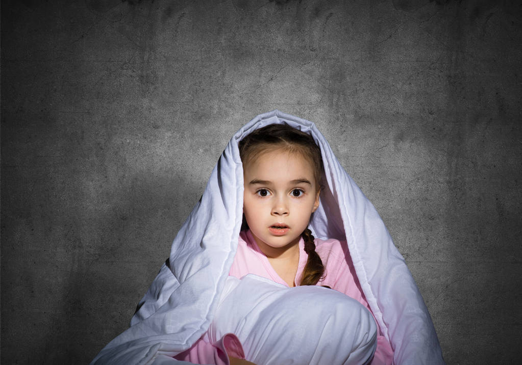 Frightened girl hiding under blanket. Scared kid lying in his bed at home. Fear of the dark. Beautiful little girl can not sleep at night. Portrait of child in pajamas on background of grey wall. - Photo, Image