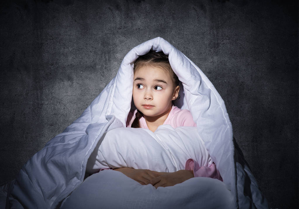 Налякана дівчина ховається під ковдрою. Налякана дитина лежить у ліжку вдома. Страх темряви. Красива маленька дівчинка не може спати вночі. Портрет дитини в піжамі на тлі сірої стіни
. - Фото, зображення