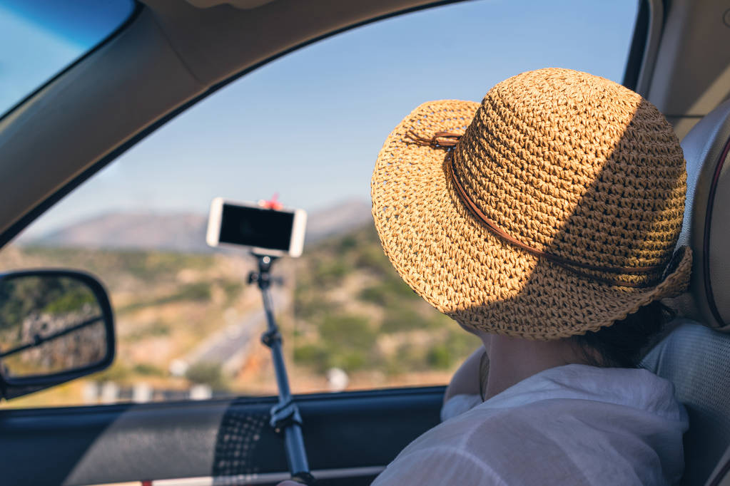 Blogger girl in the straw hat taking selfie picture or video using smartphone and selfie stick inside of car. Holidays and tourism concept - Photo, Image