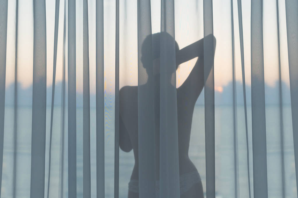 Гола красива жінка в мереживних трусиках стоїть за прозорою завісою на скляному балконі рано вранці на світанку. Вид на море
. - Фото, зображення