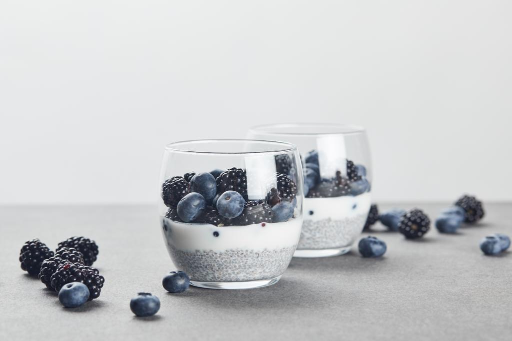 selective focus of tasty yogurt with chia seeds, blueberries and blackberries in glasses near scattered berries on marble surface isolated on grey - Photo, Image