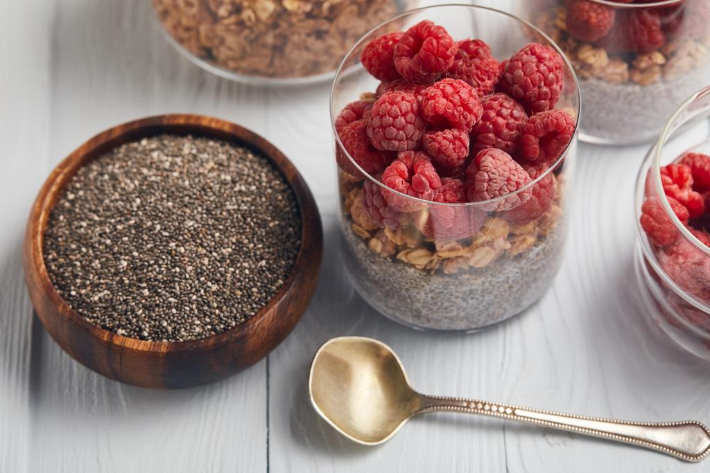 glasses with yogurt, oat flakes and raspberries near wooden bowl with chia seeds on white table  - Photo, Image