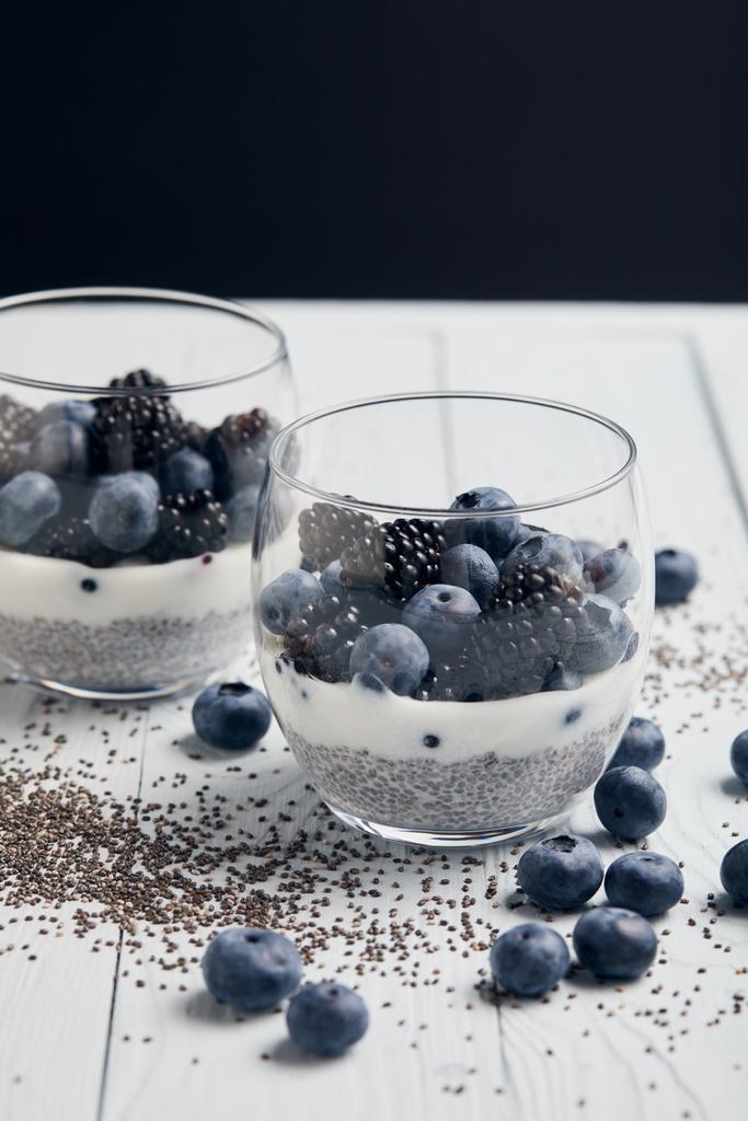 селективный фокус вкусного йогурта с семенами чиа, черникой, ежевикой рядом с разбросанными семенами и ягодами на белом деревянном изолированном на черном
 - Фото, изображение