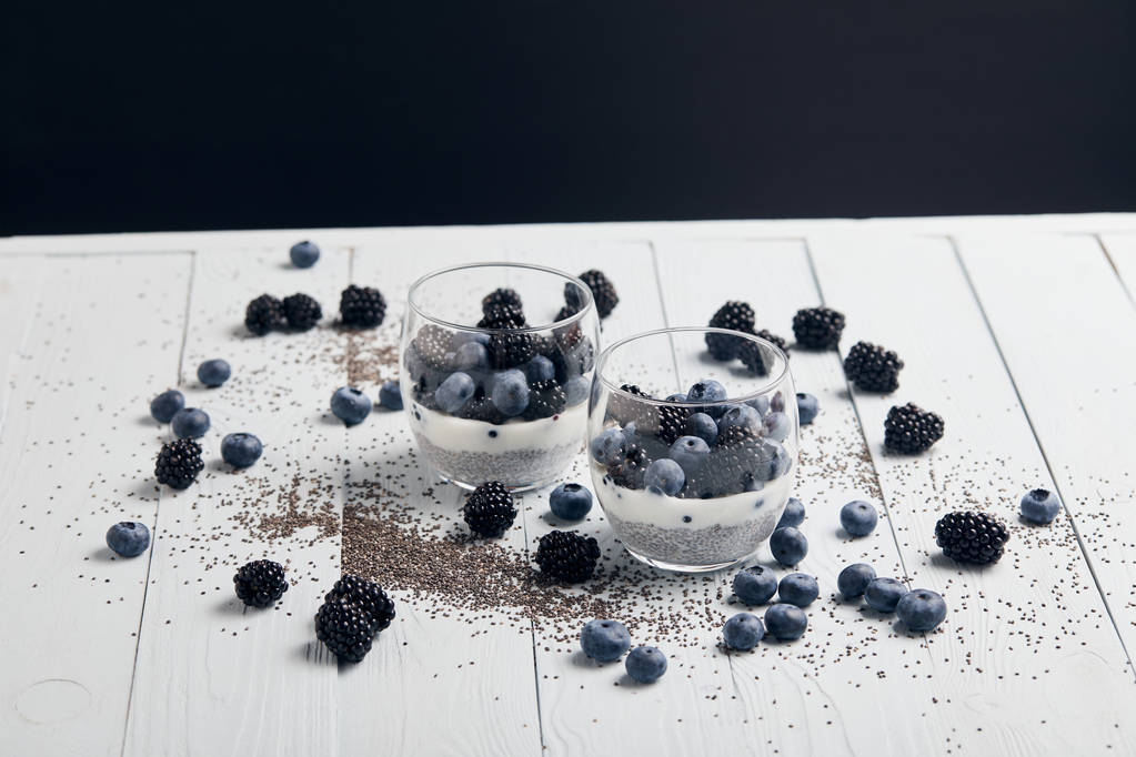 smaczny jogurt z nasion Chia, jagody, jeżyny w pobliżu rozproszonych nasion i jagód na białym drewnie na czarno - Zdjęcie, obraz