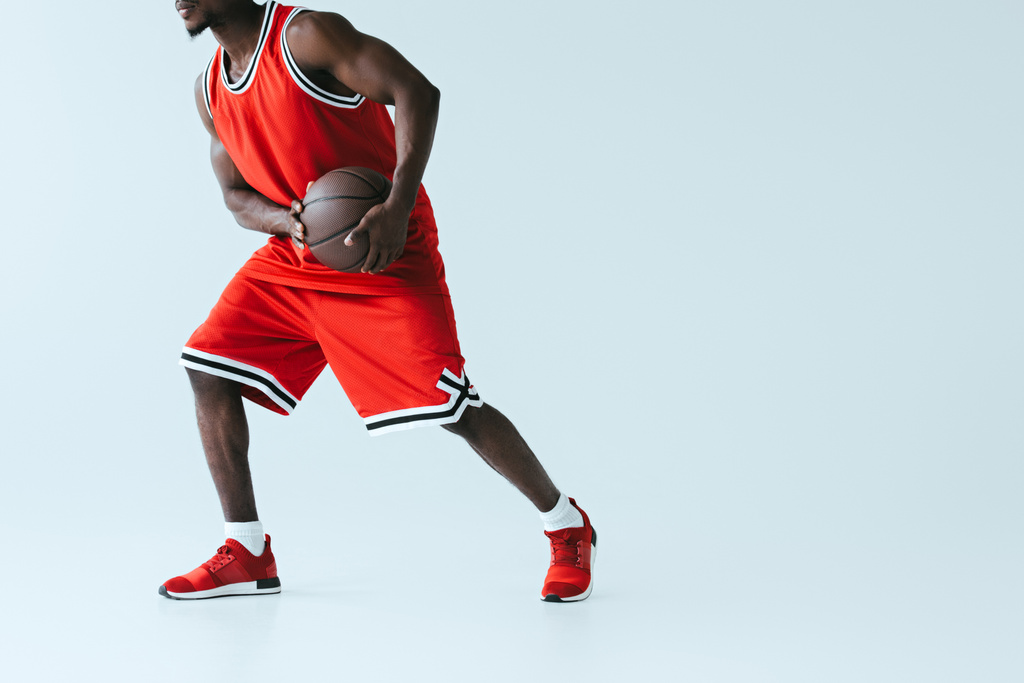 gedeeltelijke weergave van African American sportman in Red Sportswear basketbal op grijze achtergrond spelen - Foto, afbeelding