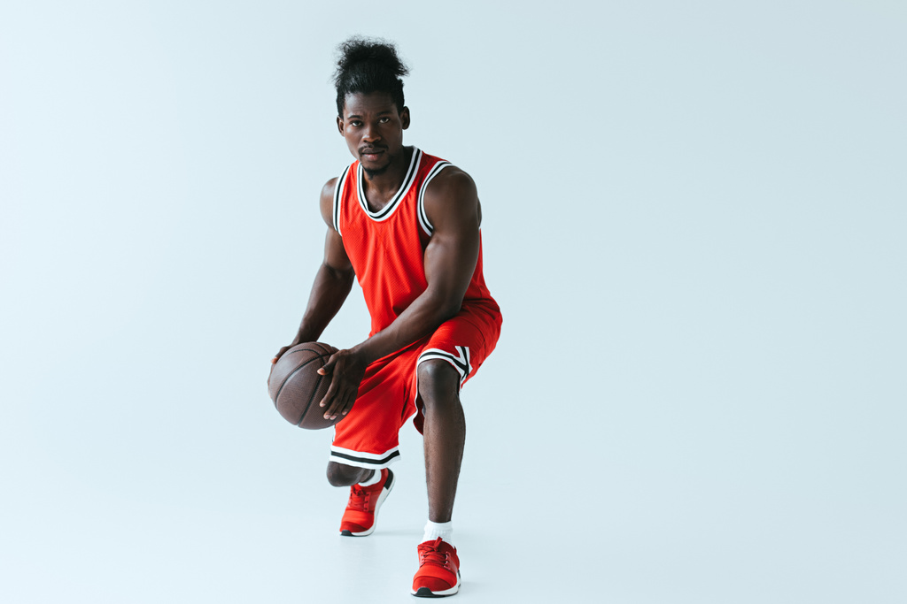 pohledný africký americký sportovec v červeném sportovním oblečení hrát basketbal na šedém pozadí - Fotografie, Obrázek