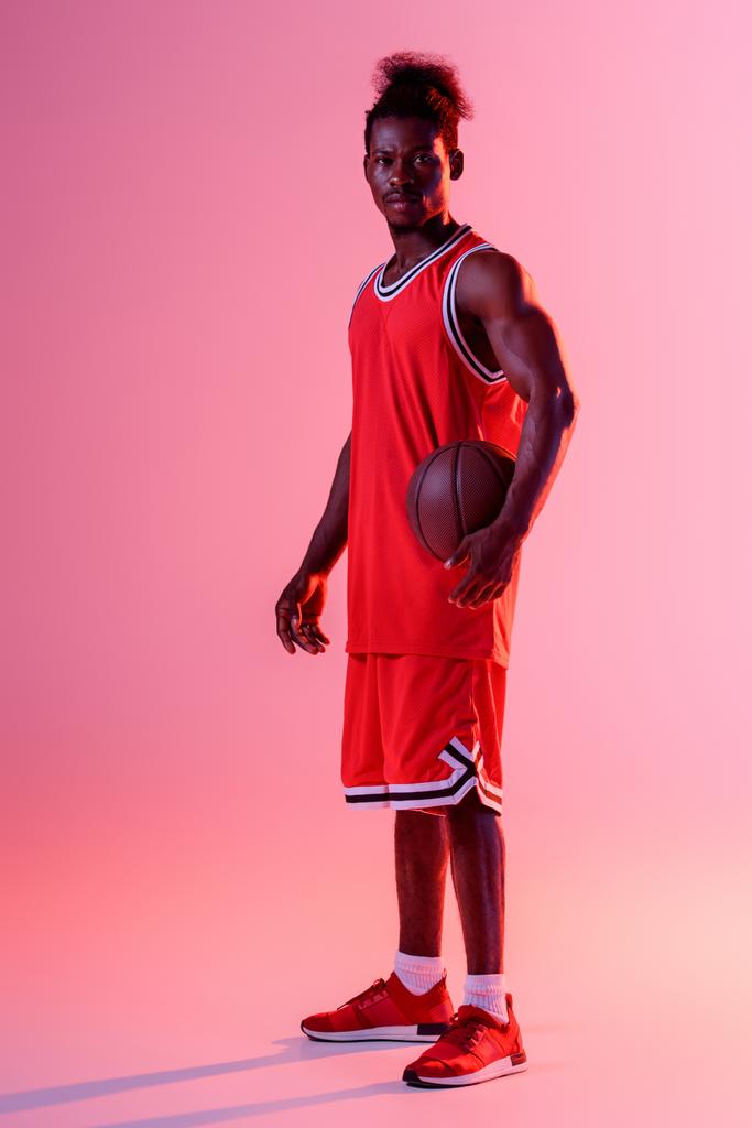 vakava afrikkalainen amerikkalainen koripalloilija katselee kameraa pitäen palloa vaaleanpunaisella taustalla valaistuksella ja kaltevuudella
 - Valokuva, kuva
