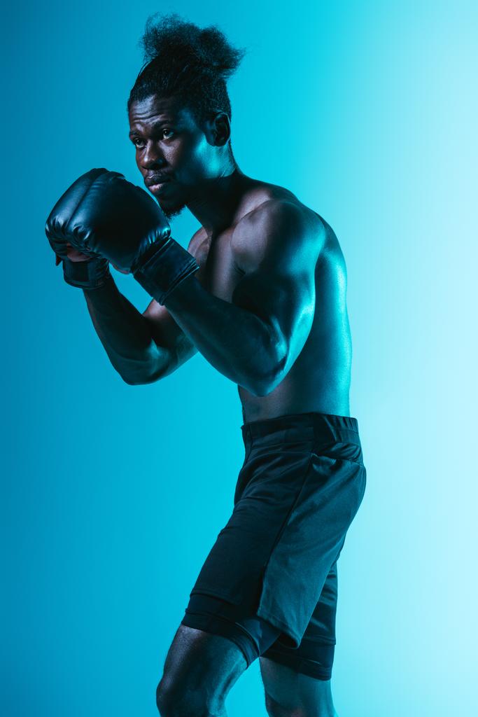 selbstbewusster afrikanisch-amerikanischer Sportler mit muskulösem Oberkörper beim Boxen auf blauem Hintergrund - Foto, Bild