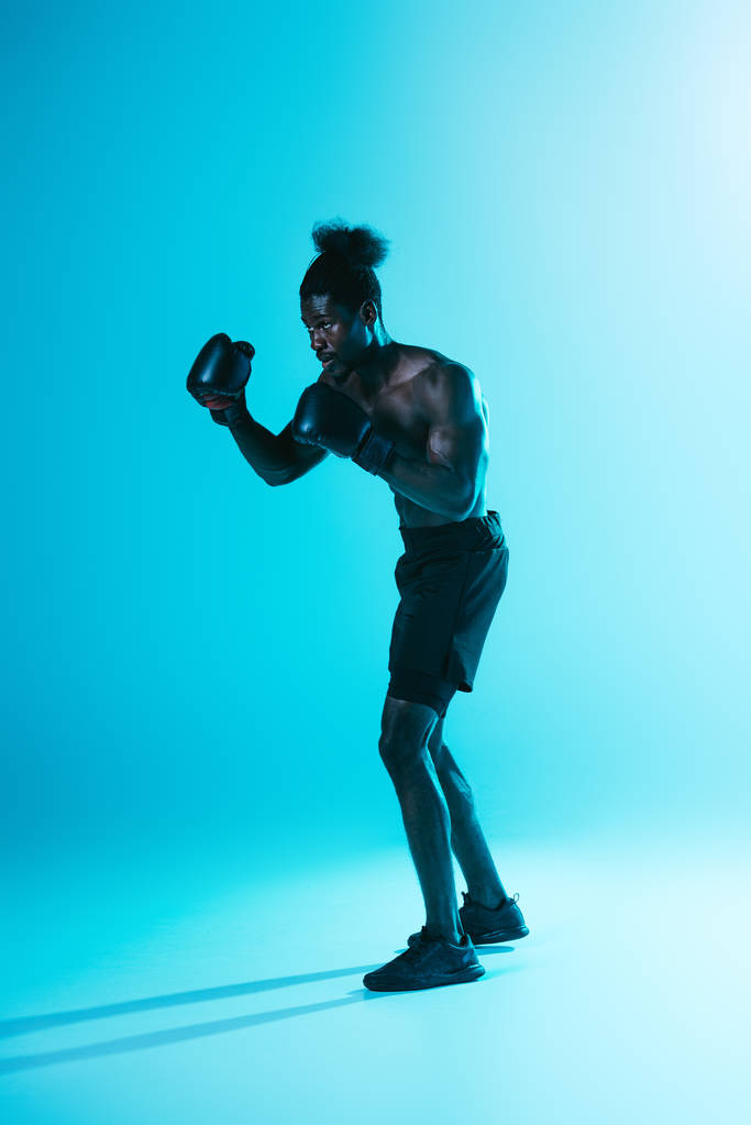 serieuze Afro-Amerikaanse sportman in shorts en sneakers Boksen op blauwe achtergrond - Foto, afbeelding