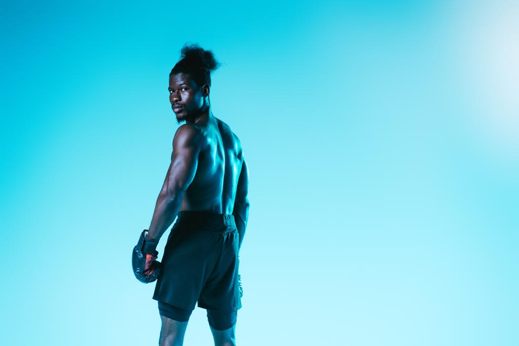 Selbstbewusster afrikanisch-amerikanischer Sportler in Boxhandschuhen blickt in die Kamera auf blauem Hintergrund - Foto, Bild