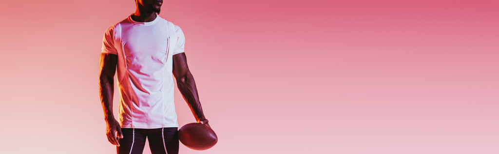 vista parziale dello sportivo afroamericano con palla da rugby su sfondo rosa con pendenza, tiro panoramico
 - Foto, immagini