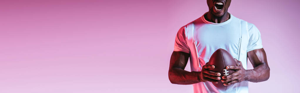 グラデーション、パノラマショットで紫色の背景にラグビーボールを保持しながら叫ぶアフリカ系アメリカ人のスポーツマンの部分的なビュー - 写真・画像
