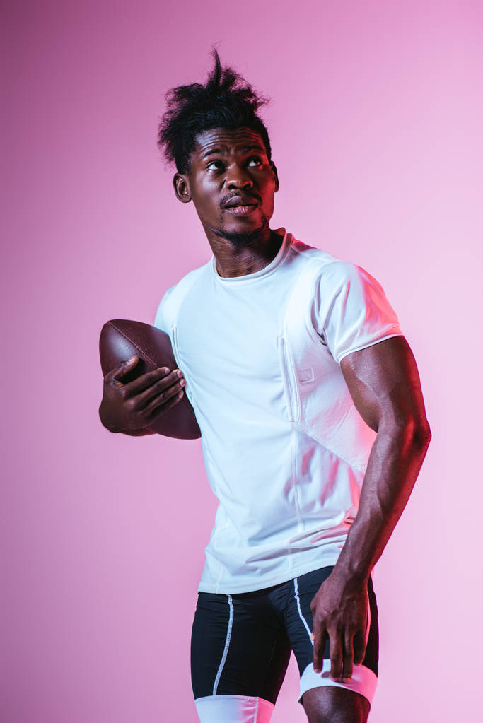 apuesto deportista afroamericano mirando hacia otro lado mientras sostiene la pelota de rugby sobre fondo púrpura con gradiente
 - Foto, Imagen