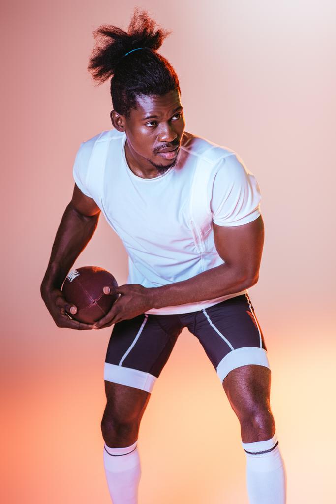 グラデーションと黄色の照明でピンクの背景にアメリカンフットボールをプレイする自信のあるアフリカ系アメリカ人のスポーツマン - 写真・画像