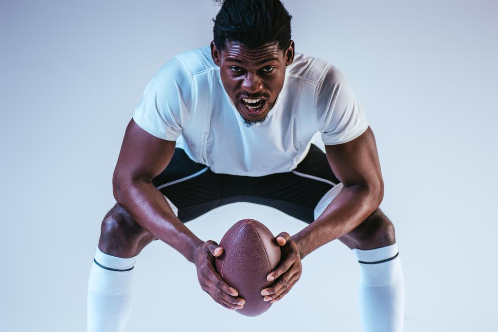 ενθουσιασμένος Αφρικανός Αμερικανός αθλητής ουρλιάζοντας ενώ παίζοντας αμερικανικό ποδόσφαιρο σε μπλε φόντο - Φωτογραφία, εικόνα