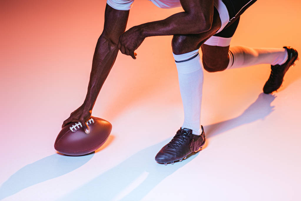 rajattu näkymä afrikkalainen amerikkalainen urheilija pelaa amerikkalaista jalkapalloa taustalla vaaleanpunainen ja keltainen kaltevuus ja valaistus
 - Valokuva, kuva