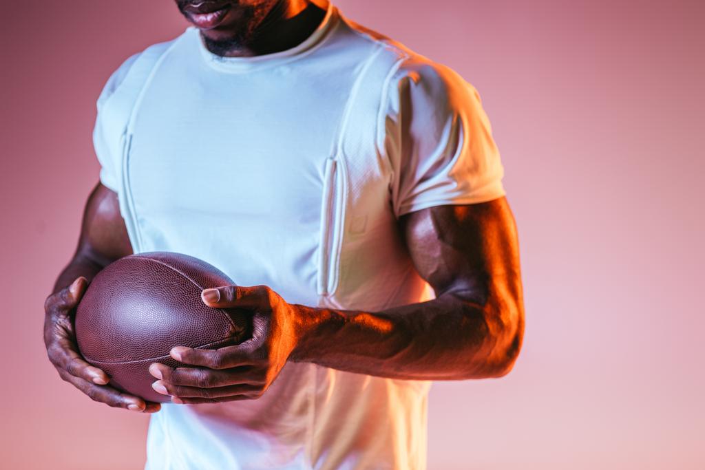 обрезанный вид афро-американского спортсмена, держащего регбийный мяч на розовом фоне с градиентом и освещением
 - Фото, изображение
