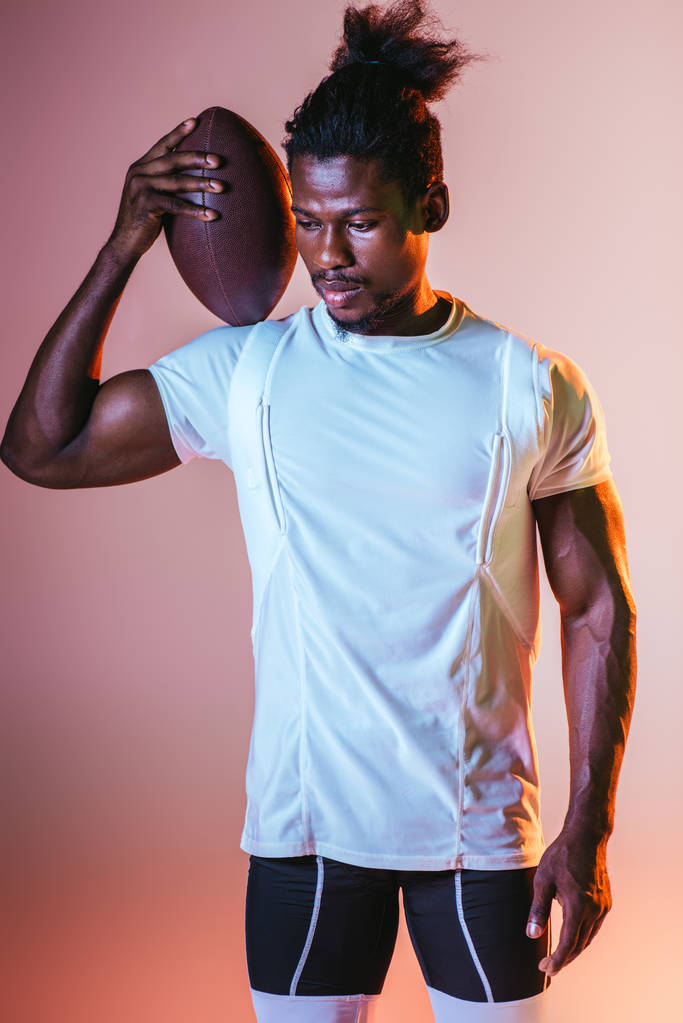 young african american sportsman holding rugby ball on pink background with gradient and lighting - Photo, Image
