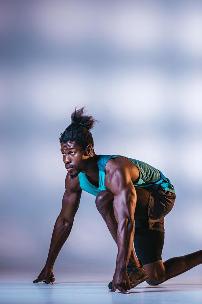 афроамериканський спортсмен, що стоїть на стартовій позиції на сірому фоні з освітленням
 - Фото, зображення