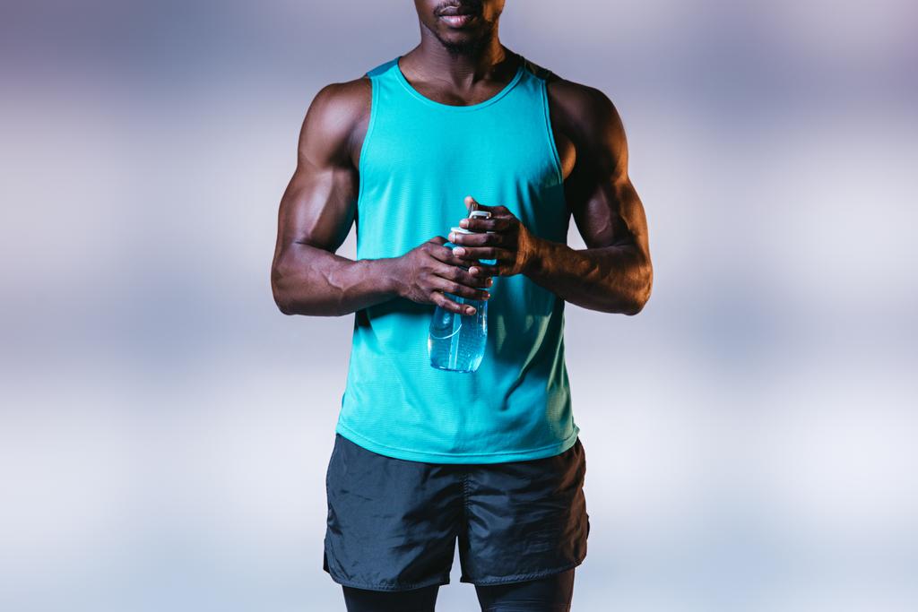 vista parcial del deportista afroamericano musculoso sosteniendo botella deportiva sobre fondo gris con iluminación
 - Foto, imagen