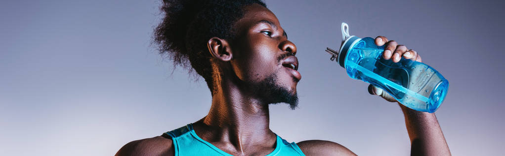 plano panorámico del deportista afroamericano bebiendo de la botella de deportes sobre fondo gris y azul degradado con iluminación
 - Foto, imagen