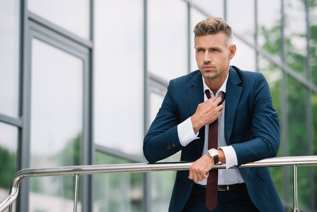 handsome businessman in suit standing outside and touching tie near building  - Photo, Image