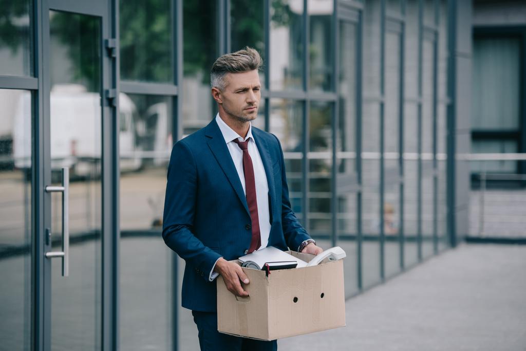homem de negócios triste e demitido de pé perto do edifício e segurando caixa de papelão
  - Foto, Imagem