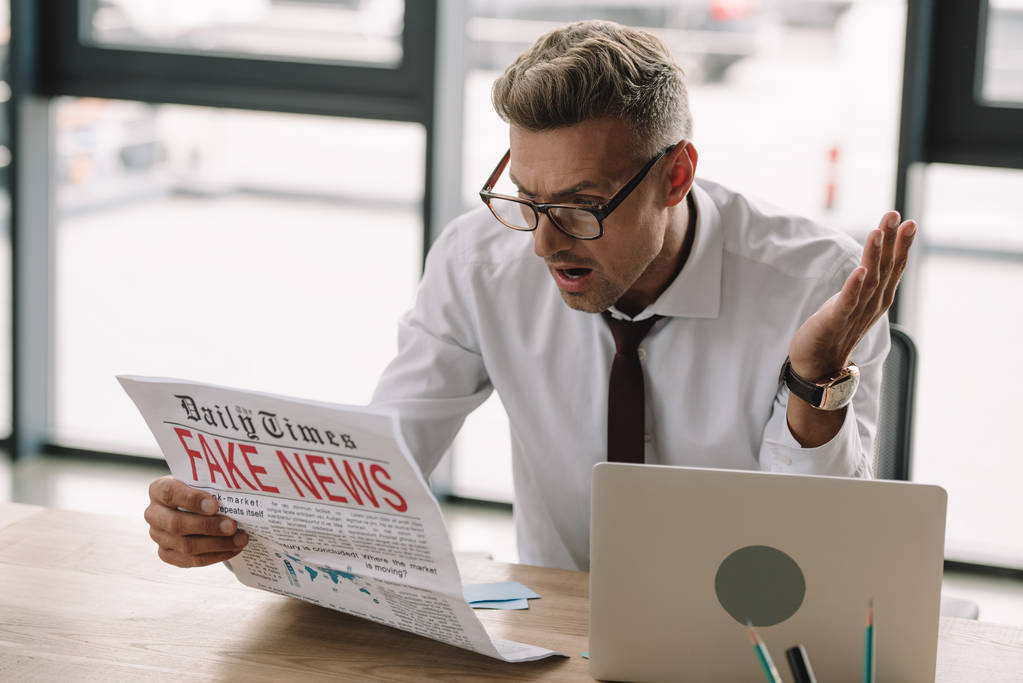 шокированный бизнесмен в очках жестикулирует и читает газету с фейковыми новостями
  - Фото, изображение