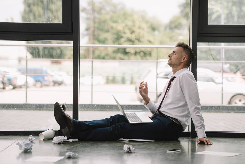 πλευρική όψη του όμορφου επιχειρηματία με κλειστά μάτια που κάθονται στο πάτωμα με φορητό υπολογιστή  - Φωτογραφία, εικόνα