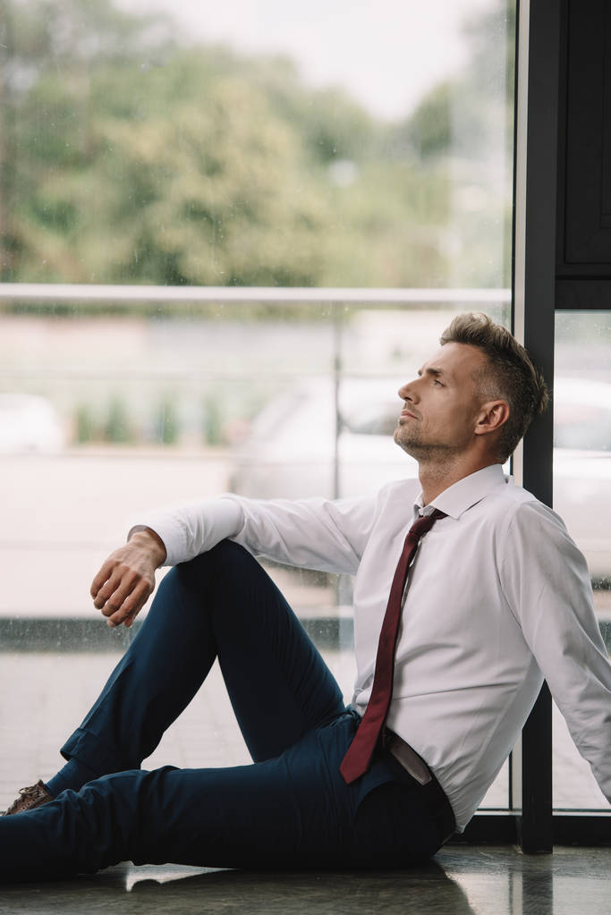 オフィスの窓の近くの床に座っているハンサムなビジネスマン  - 写真・画像