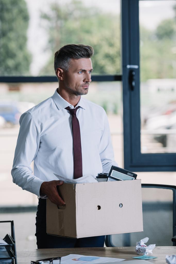 όμορφος επιχειρηματίας σε κοστούμι κρατώντας κουτί κοντά στο χώρο εργασίας  - Φωτογραφία, εικόνα