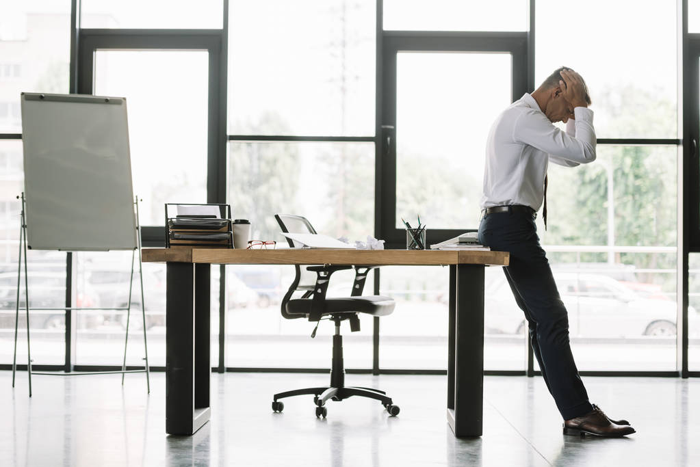 guapo hombre de negocios tocando la cabeza mientras está de pie cerca de la mesa en la oficina moderna
  - Foto, imagen