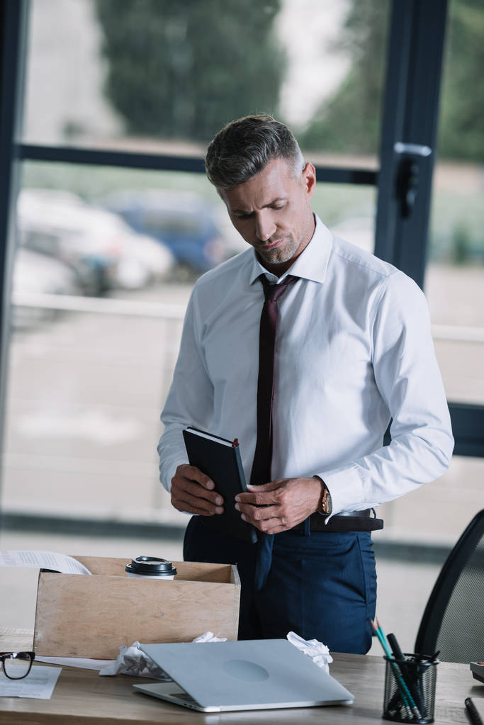 displeased businessman holding notebook near box on table  - Photo, Image