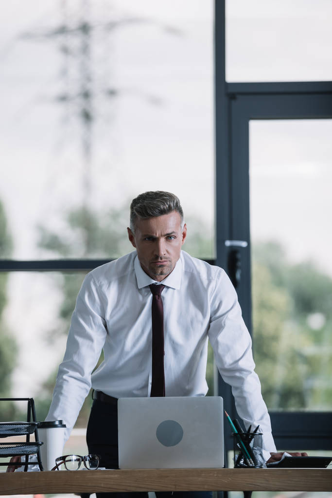 serious businessman in formal wear standing near desk and looking at camera  - Photo, Image