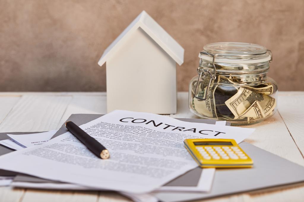 модель дома на белом деревянном столе с контрактом, калькулятор и деньги возле коричневой текстурированной стены, концепция недвижимости
 - Фото, изображение