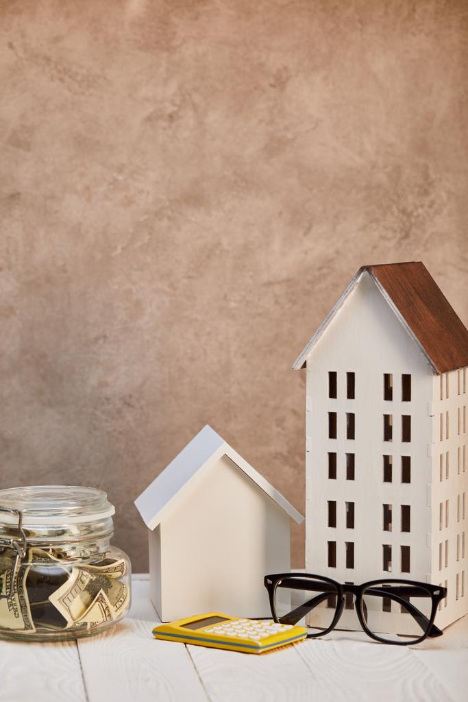 házak modellek fehér fából készült asztal moneybox, számológép és szemüveg közelében barna textúrájú fal, ingatlan koncepció - Fotó, kép