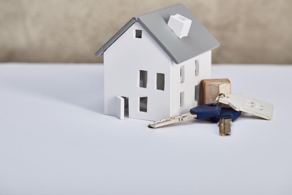 Hausmodell auf weißem Tisch mit Schlüsseln in der Nähe der grauen strukturierten Wand, Immobilienkonzept - Foto, Bild