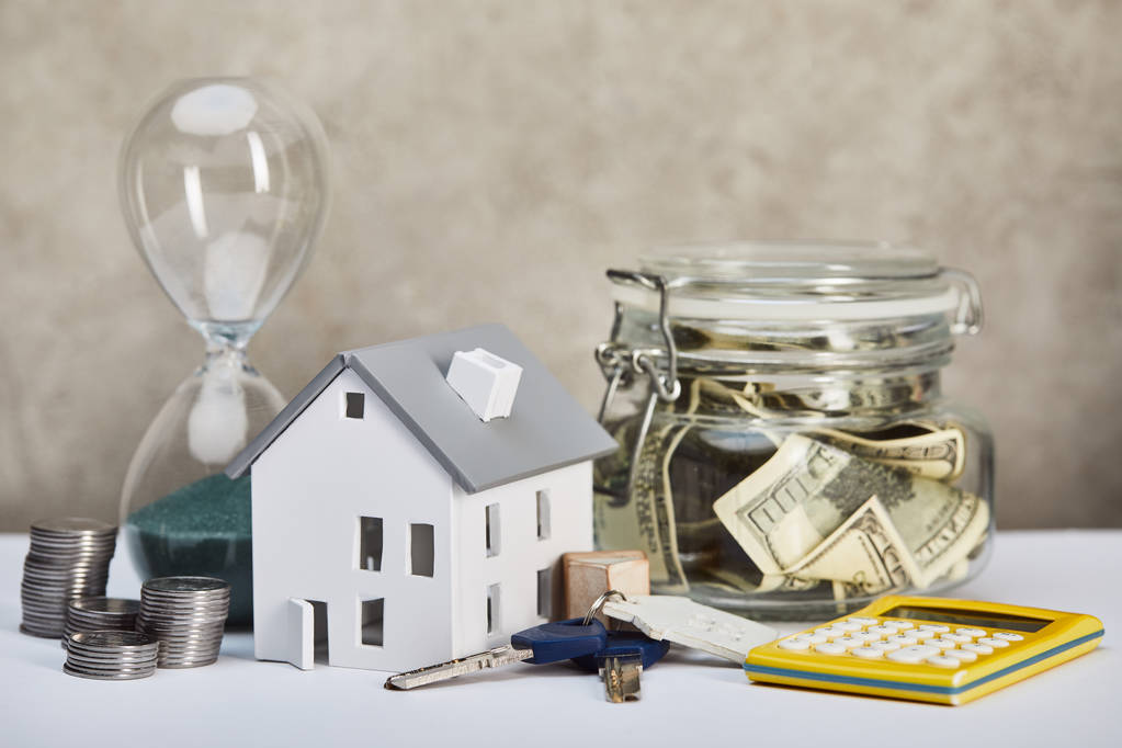model domu na bílém stole s přesýpací hodiny, kalkulačka, peníze a klíče, koncepce nemovitostí - Fotografie, Obrázek
