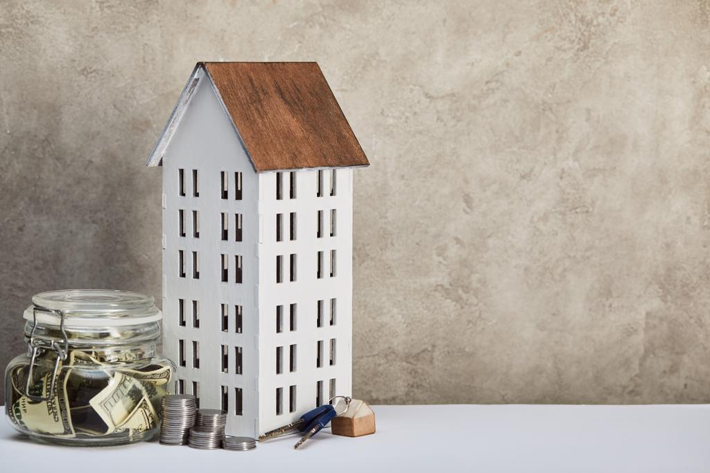 Hausmodell, Spardose mit Dollarnoten, Schlüsseln und Silbermünzen auf weißem Tisch auf grauem Hintergrund, Immobilienkonzept - Foto, Bild