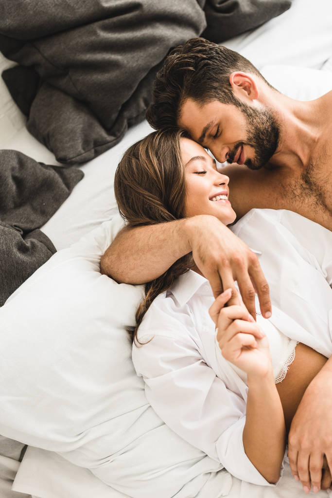 Szexi fiatal pár az ágyban fekve, mosolyogva csukott szemmel, átölelve és kezében kezek - Fotó, kép