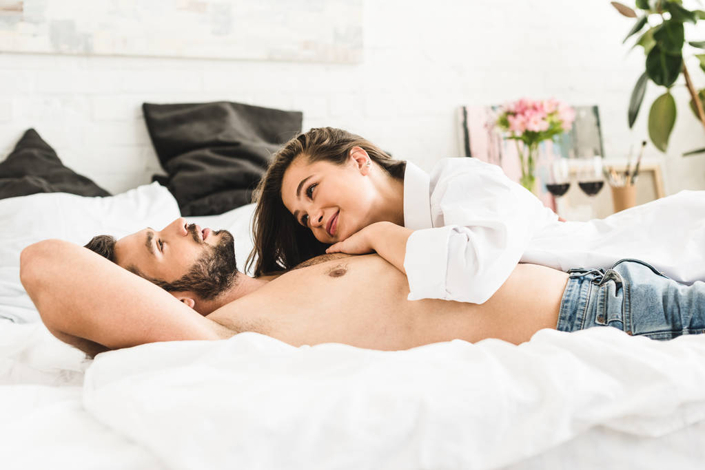 女の子が微笑んで男を見ている間、若いカップルがベッドに横たわっている - 写真・画像