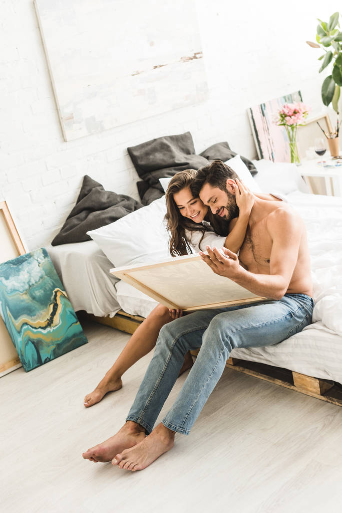 молодая пара сидит в постели и улыбается, в то время как мужчина держит картину и девушка обнимает парня
 - Фото, изображение