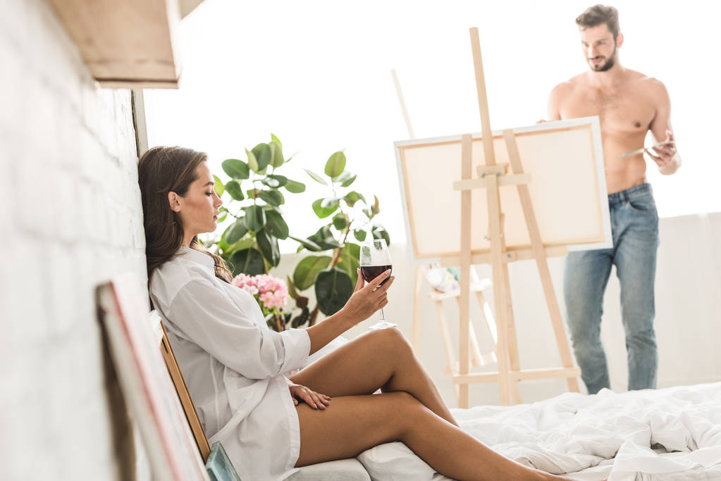 vista lateral de la chica sentada en la cama con vino mientras el hombre sin camisa dibujo novia con pincel y pinturas de colores
 - Foto, Imagen