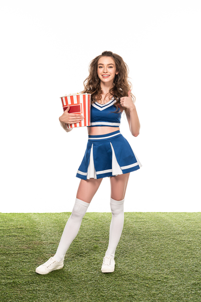 щаслива дівчина-вболівальник у блакитній формі, що стоїть з відром попкорну і показує великий палець на зеленому полі ізольовано на білому
 - Фото, зображення