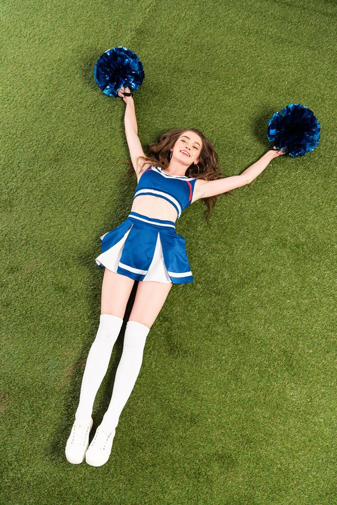 bovenaanzicht van mooie cheerleader meisje in blauw uniform liggend met pompoms op groen veld - Foto, afbeelding